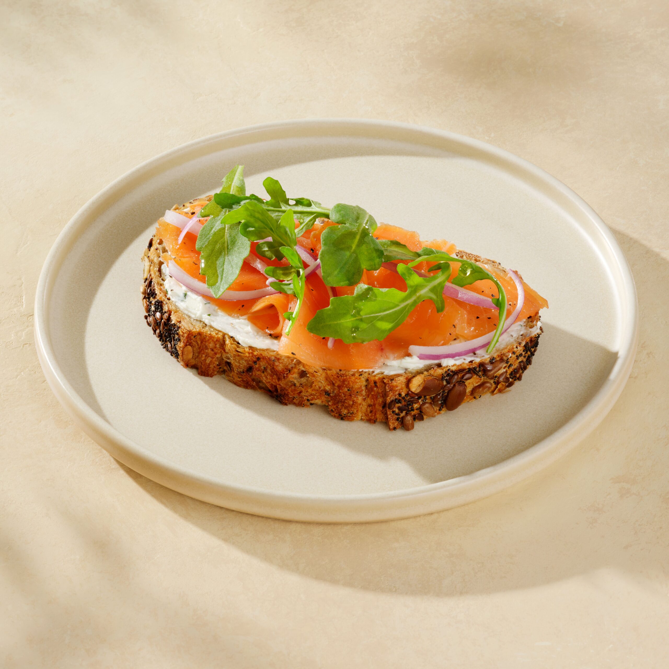 A slice of multigrain bread topped with cream cheese, smoked salmon, red onion, and fresh arugula, served on a light plate.