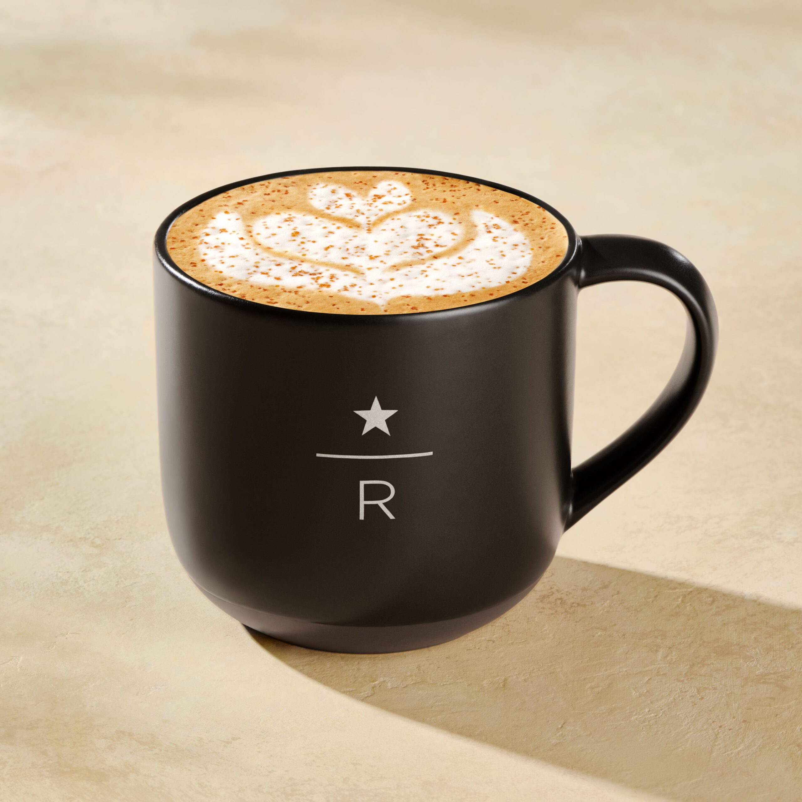 A black coffee mug with a smooth finish features a latte topped with a decorative foam design and a sprinkle of cinnamon.