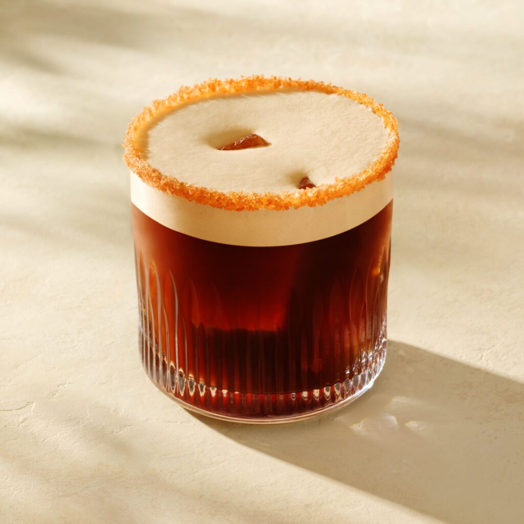 A glass of dark beverage topped with a creamy layer and a sugared rim. The glass has a textured design and sits on a light surface.
