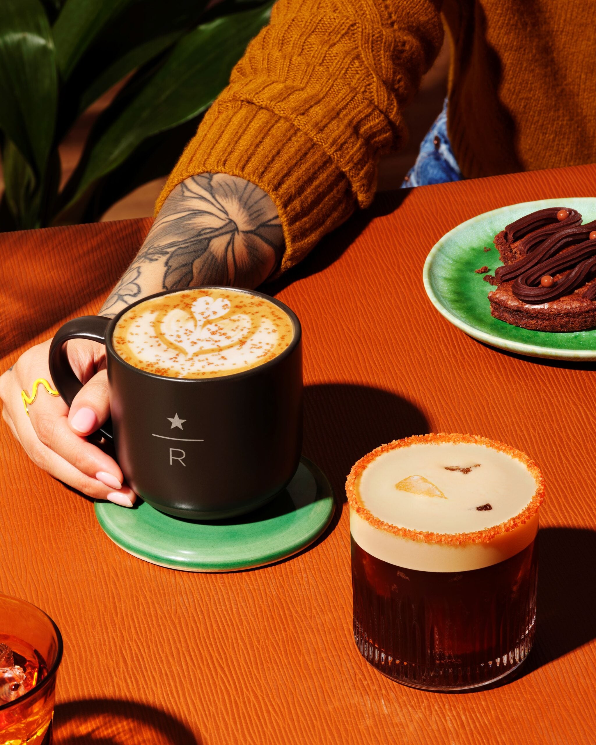 A hand holds a dark coffee mug with latte art, next to a glass of a layered drink with a frothy top and a sugared rim. A plate with chocolate dessert is also visible. The setting features a textured orange table and green accents.