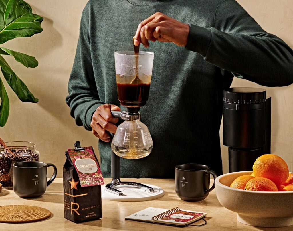A person stirs coffee in a siphon brewer. Nearby are a bag of Costa Rican coffee, a black mug, a notebook, and a bowl of oranges.