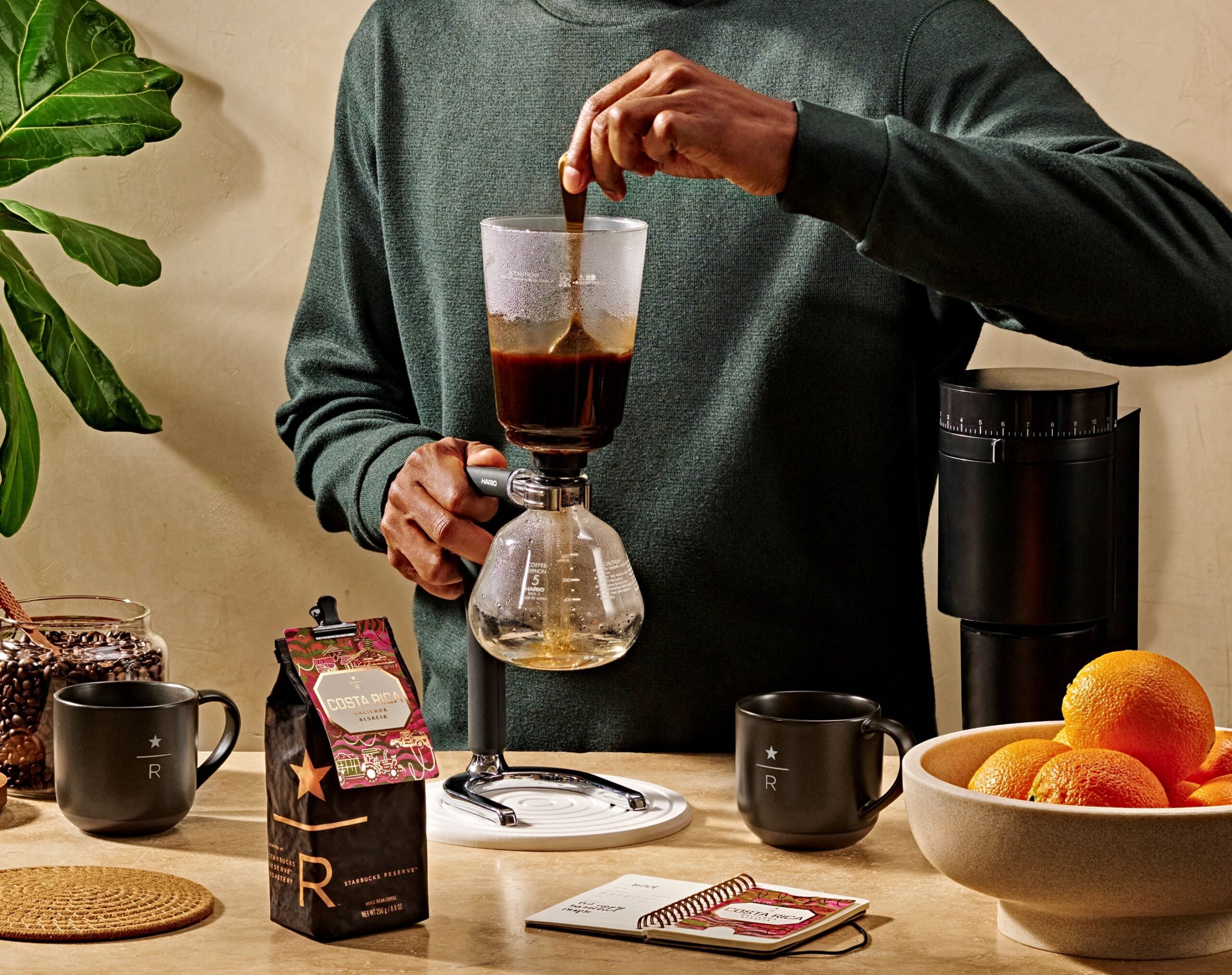 A person stirs coffee in a siphon brewer. Nearby are a bag of Costa Rican coffee, a black mug, a notebook, and a bowl of oranges.