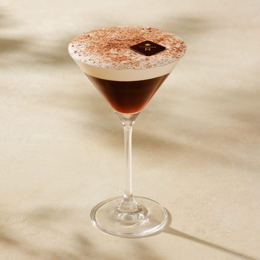 A sleek martini glass filled with dark coffee, topped with a frothy layer and a dusting of cocoa powder, featuring a chocolate square.