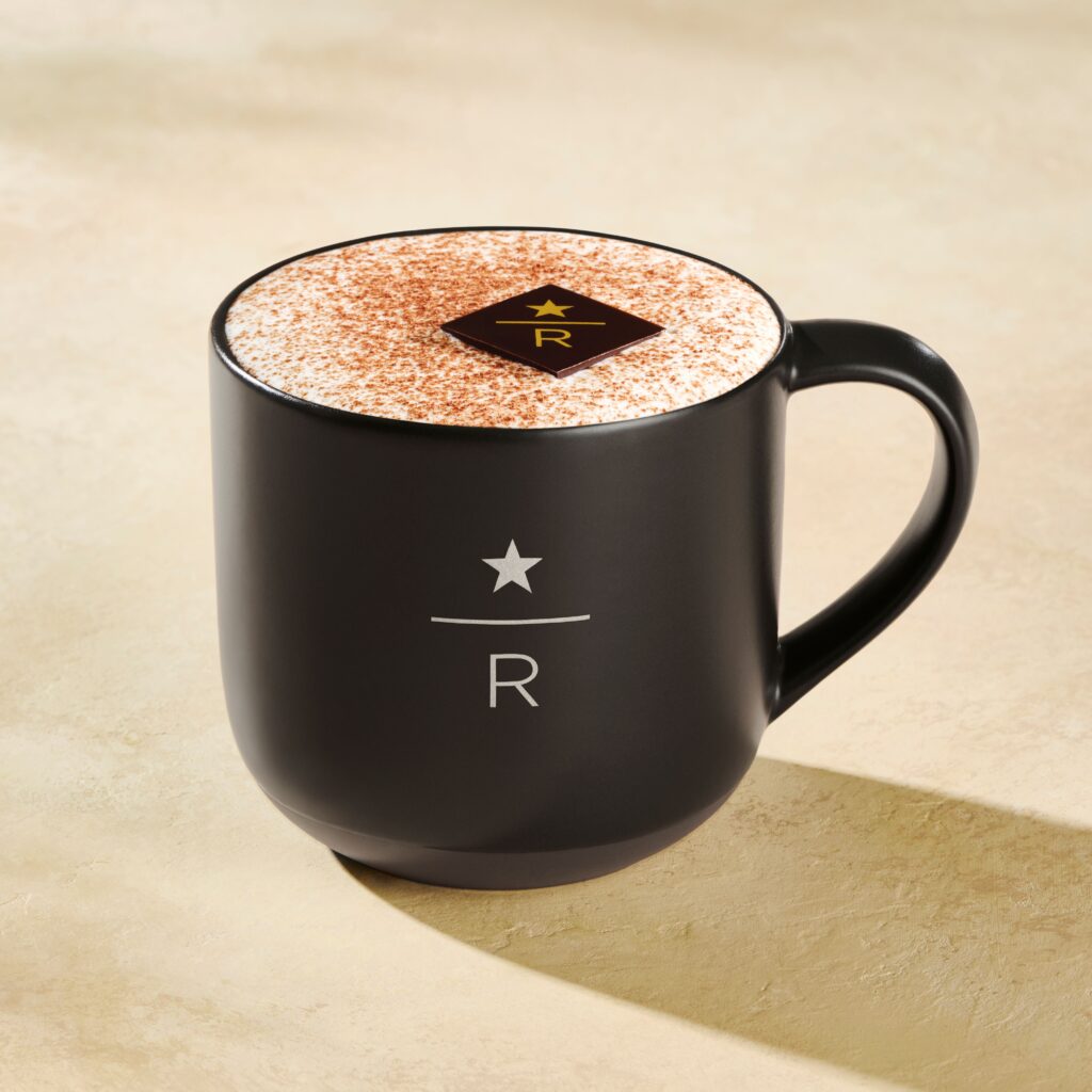 A black coffee mug with a smooth finish, topped with frothy milk and cocoa powder, features a decorative chocolate piece on top.