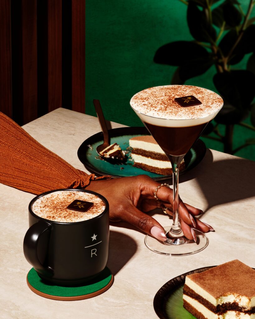 A black mug with a logo holds a frothy beverage topped with cocoa. Nearby, a martini glass contains a similar drink. Two plates feature layered desserts. A hand with manicured nails reaches for the martini. The background is a vibrant green.