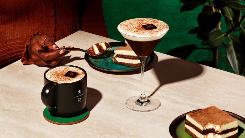 A close-up of a table setting featuring a martini glass with a creamy coffee drink topped with cocoa powder, a black mug with a logo, and a plate of layered cake. A hand is holding a fork, ready to take a bite of the cake. The background is a vibrant green.