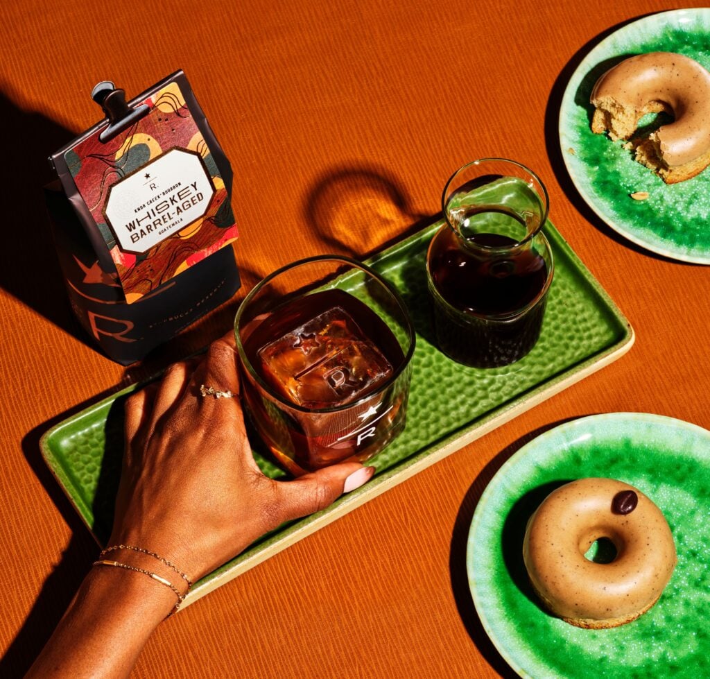 A hand holds a glass with whiskey and ice, accompanied by a small carafe of dark liquid on a green tray. Nearby, two plates feature a donut with a bite taken out and a whole donut, both on green dishes. The background is a warm orange.