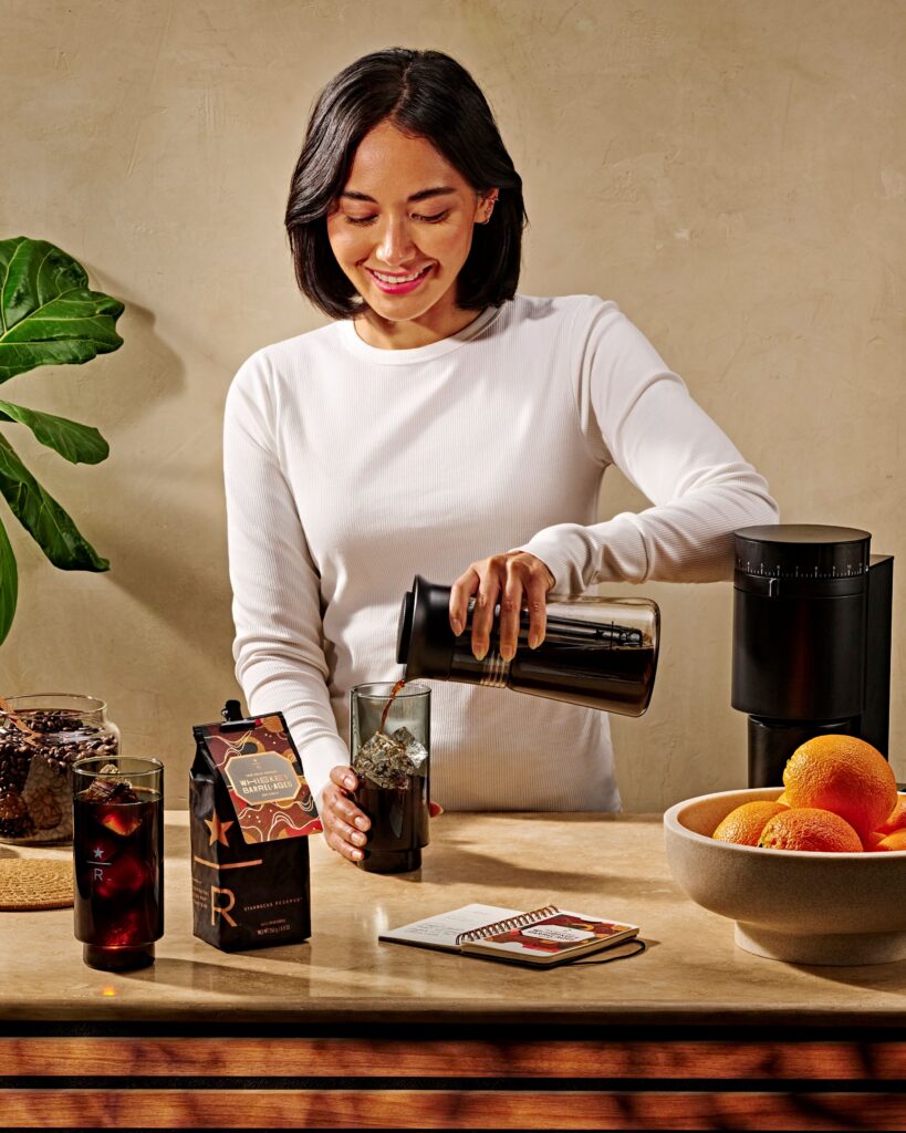 A woman in a white long-sleeve shirt pours coffee from a carafe into a glass with ice. A carton of coffee, a bowl of oranges, and a notebook are on the table.