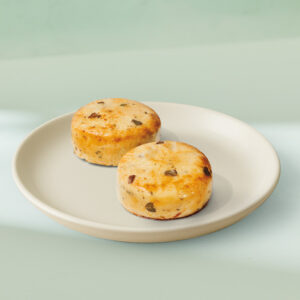 Two round, golden-brown baked goods with a slightly crispy top, placed on a light-colored plate. They appear to have bits of dark ingredients mixed in.