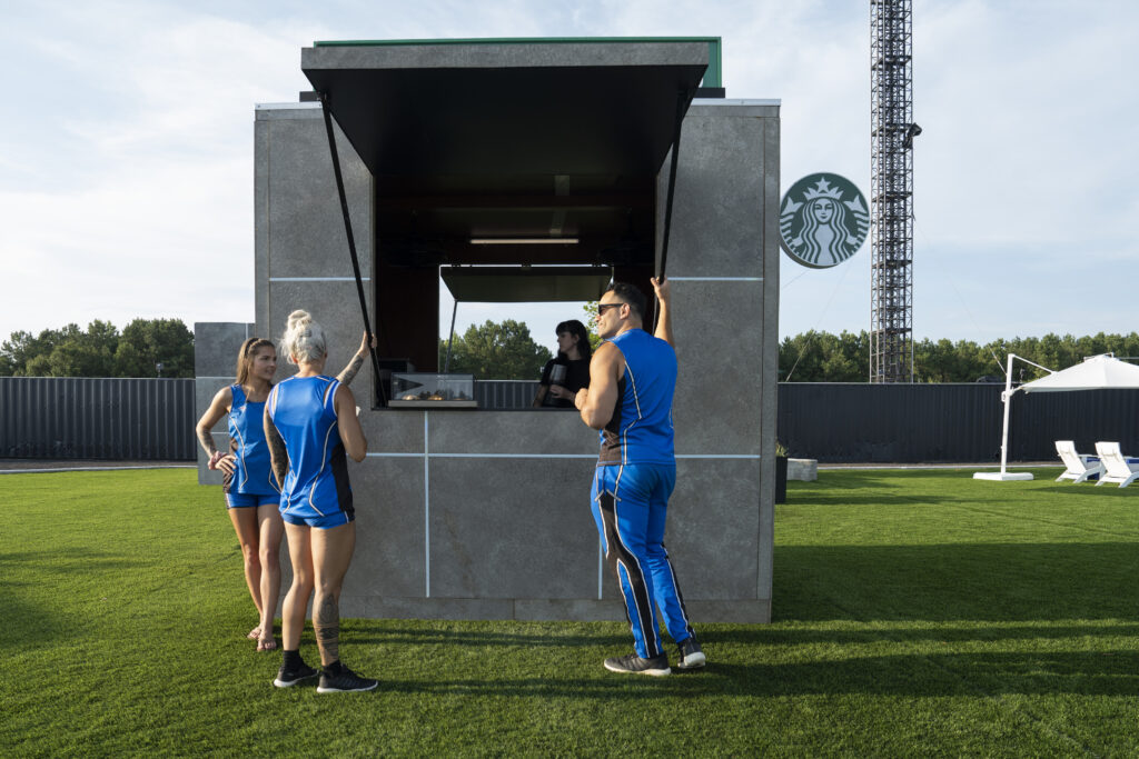 A modern outdoor kiosk with a serving window is shown. Three individuals in blue athletic outfits are interacting with a barista inside. The setting features green grass and a Starbucks logo in the background.