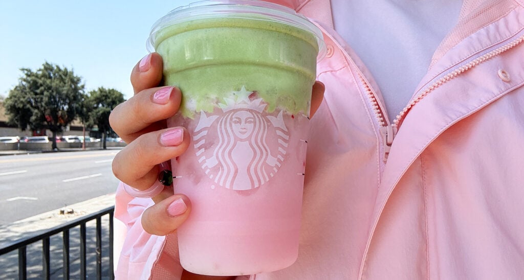 A hand holds a Starbucks cup featuring a layered drink with pink and green colors. The person is wearing a light pink jacket.