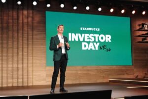 A speaker in a green blazer stands on stage, presenting at Starbucks' Investor Day event. A large screen behind displays "STARBUCKS INVESTOR DAY NYC '26" in bold white text against a green background. The stage features wooden paneling and warm lighting.