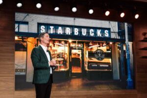 A man in a green blazer stands in front of a large screen displaying a Starbucks storefront, featuring its iconic logo and signage.