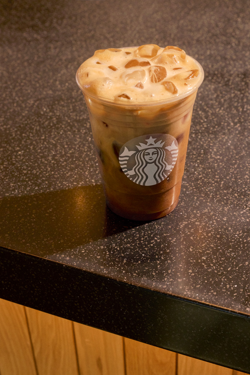A clear cup filled with iced coffee topped with frothy cream sits on a dark speckled countertop.