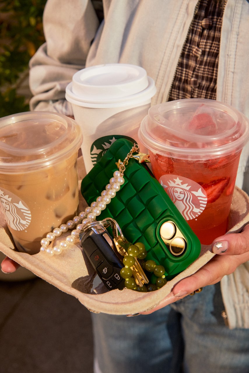 A person holds a drink carrier with three beverages: two Starbucks cups (one iced coffee and one iced drink) and a third cup with a lid. A green phone case and car keys are also visible, along with a beaded keychain.