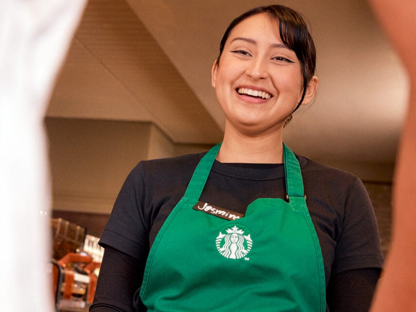 A barista in a green apron smiles while serving two white cups to customers. The setting appears to be a coffee shop.
