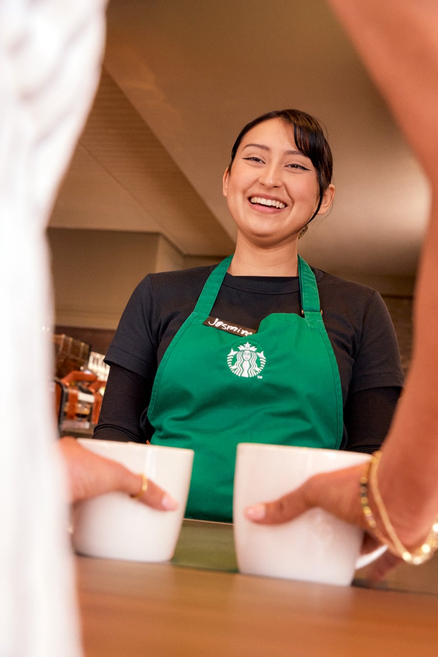 A barista in a green apron smiles while serving two white cups to customers. The setting appears to be a coffee shop.