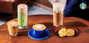 A wooden table displays five beverages: a glass of iced green tea, a tall iced coffee, a small cup of latte, a glass of chai, and two baked goods on a plate.