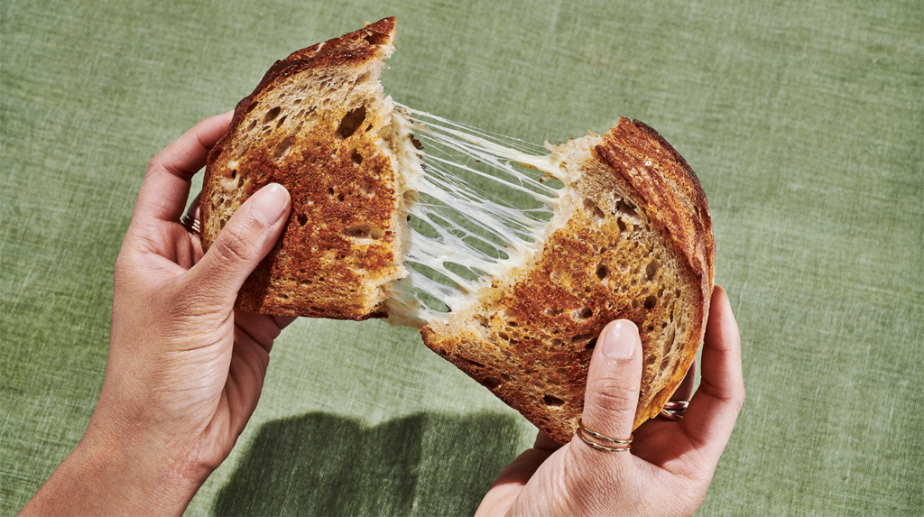 A person is pulling apart a grilled cheese sandwich, revealing gooey melted cheese stretching between two slices of golden-brown bread. The background is a textured green surface.