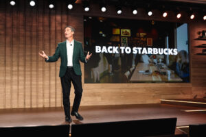 A speaker in a green blazer gestures while presenting on stage. Behind him, a large screen displays the text "BACK TO STARBUCKS" alongside an image of a Starbucks interior.