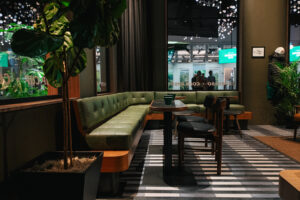 A cozy seating area features a green upholstered bench along the wall, a small table with green cups, and a few black chairs. A potted plant adds a touch of greenery, while large windows provide natural light. The floor has a striped pattern.