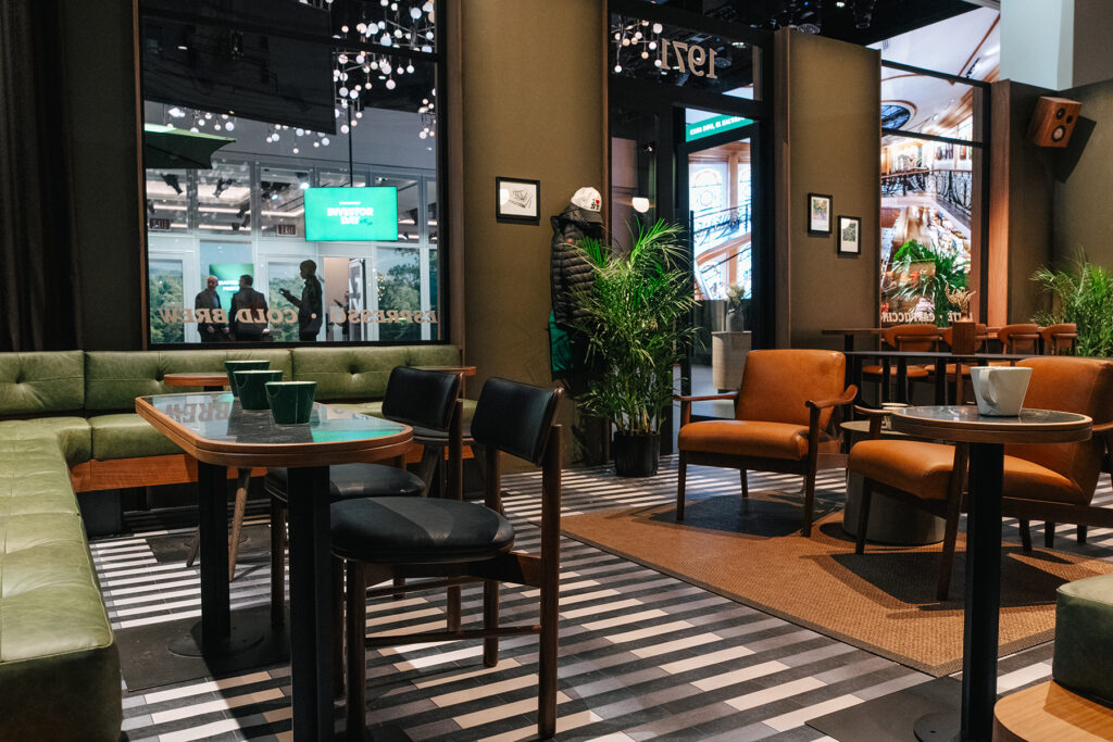 A modern café interior featuring green leather seating, wooden tables, and a mix of black and brown chairs. Potted plants add greenery, while large windows showcase a view of people outside. The floor has a striped pattern, enhancing the contemporary vibe.