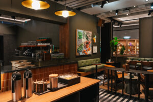 A modern café interior featuring a dark color scheme, green seating, a coffee bar with glassware, and decorative plants.
