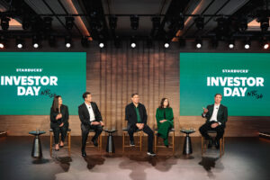 A panel of five speakers is seated on stage during Starbucks' Investor Day event. The backdrop features a large green screen with the text "STARBUCKS' INVESTOR DAY NYC '26." The speakers are dressed in business attire, with two women and three men, discussing in front of an audience.