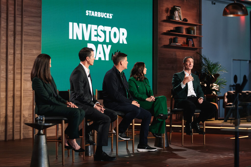 A panel of four speakers is seated on stage during Starbucks' Investor Day, with a large green backdrop displaying "INVESTOR DAY." The setting features wooden accents and plants.