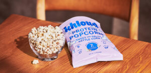 A glass bowl filled with popcorn sits on a wooden table next to a pink bag of "khloud Protein Popcorn," labeled sweet and salty kettle corn.