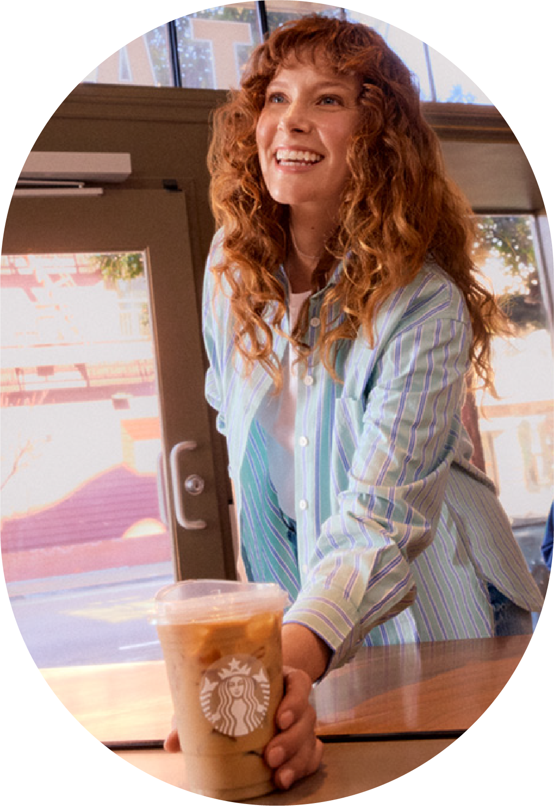A smiling woman with curly hair is reaching for a Starbucks iced drink on a table. She wears a striped shirt and is in a bright café setting.