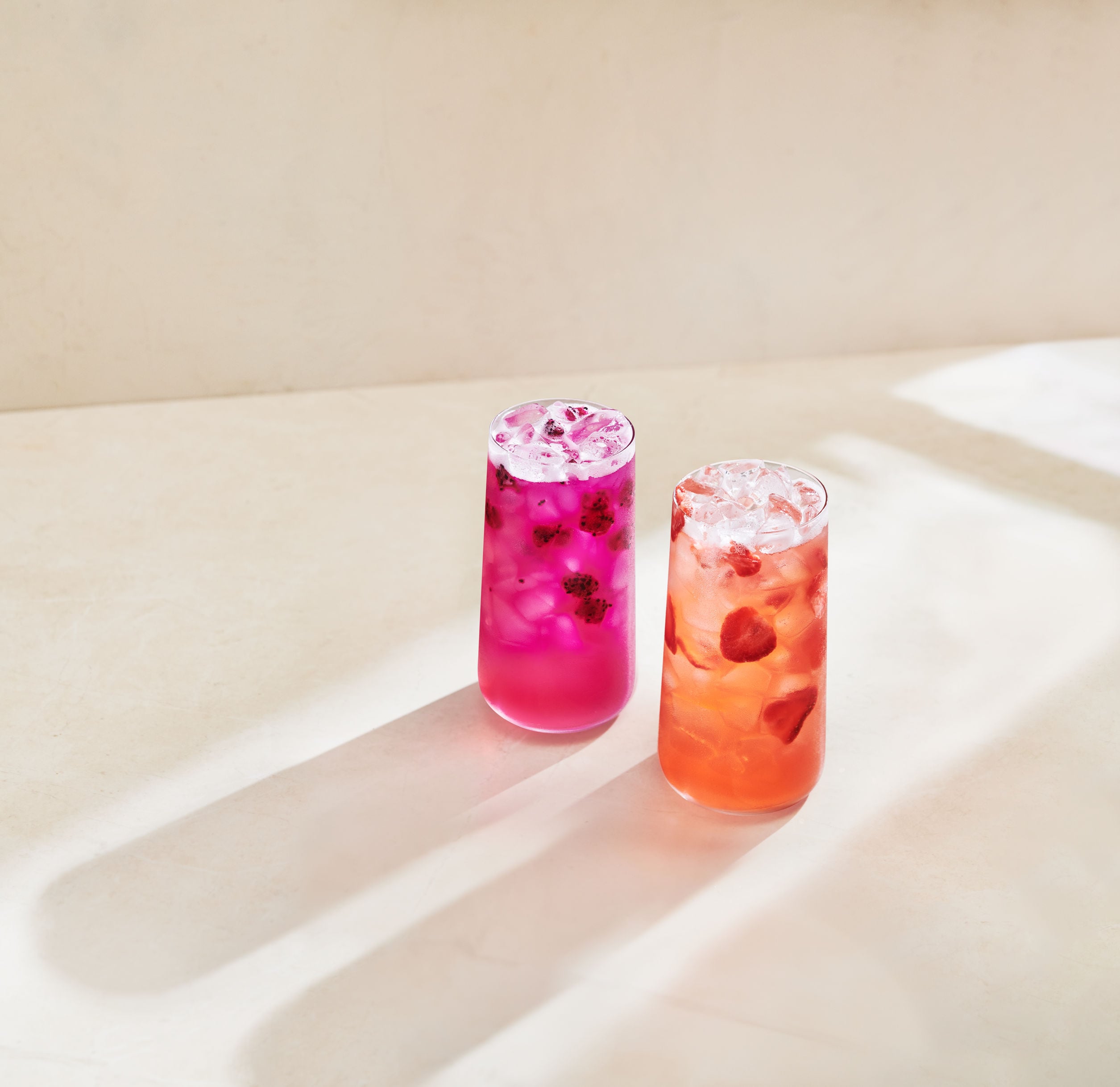 Two tall glasses of colorful drinks sit on a light surface. The left glass contains a vibrant pink beverage with ice and dark fruit pieces, while the right glass holds a lighter orange drink with ice and visible fruit slices. Soft shadows stretch across the surface.