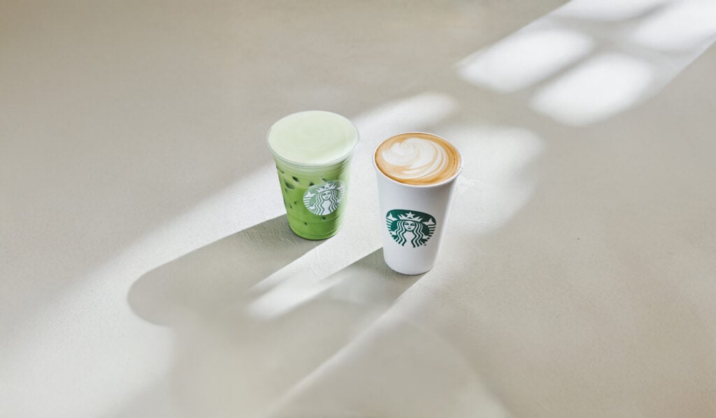 A green iced drink in a clear cup and a latte in a white cup, both featuring the Starbucks logo, are placed on a light surface.