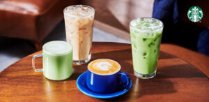 Four beverages are displayed on a wooden table: a green matcha latte in a clear mug, an iced coffee, a latte with latte art in a blue cup, and a green iced drink.