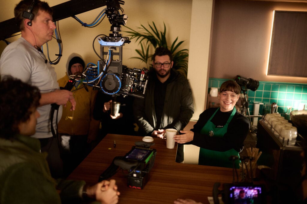 A film crew is set up in a café. A woman in a green apron is handing a cup to the camera, while crew members observe. A camera on a crane captures the scene.