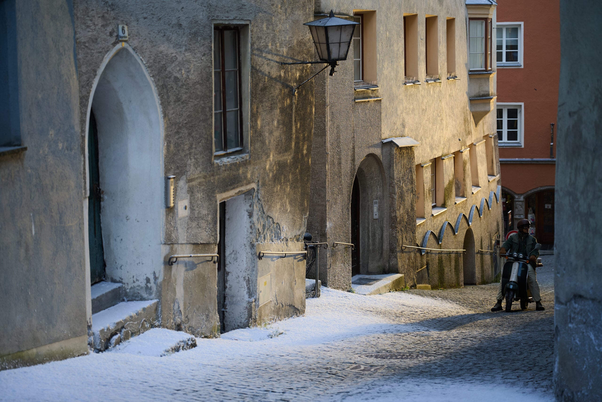 A narrow, snow-covered street features weathered buildings with textured walls, arched doorways, and a vintage lantern.