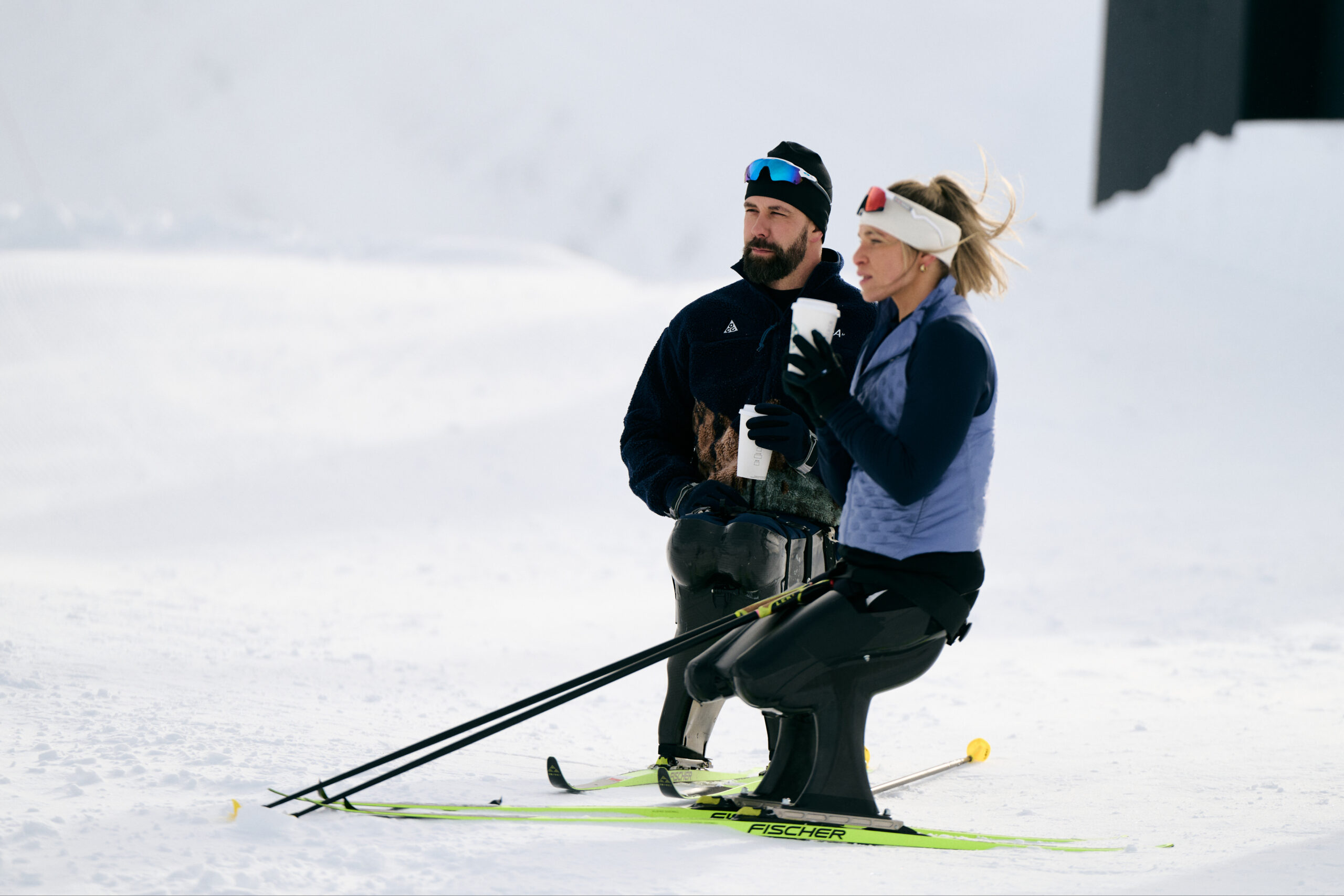 A man and a woman are on snow-covered terrain, both dressed in winter sports attire. The man holds a drink and wears a dark fleece jacket and sunglasses. The woman, seated, is in a light blue vest and has a headband. Ski poles are positioned beside them.