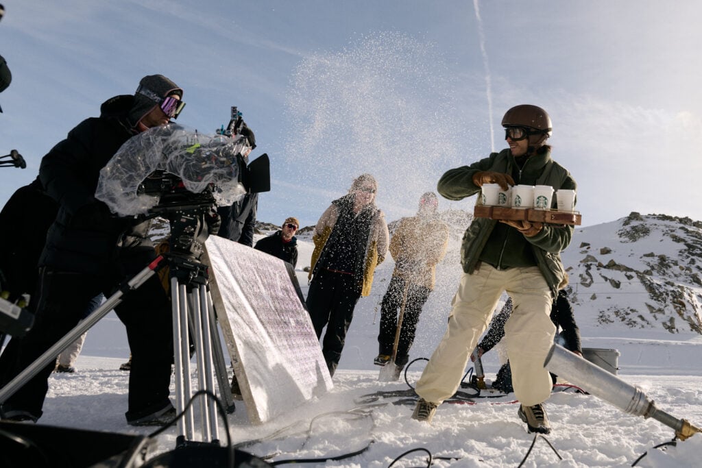 A snowy outdoor scene features a film crew with a camera and a person in a helmet carrying a tray of Starbucks cups. Snow is being kicked up around them, while others watch in winter attire.