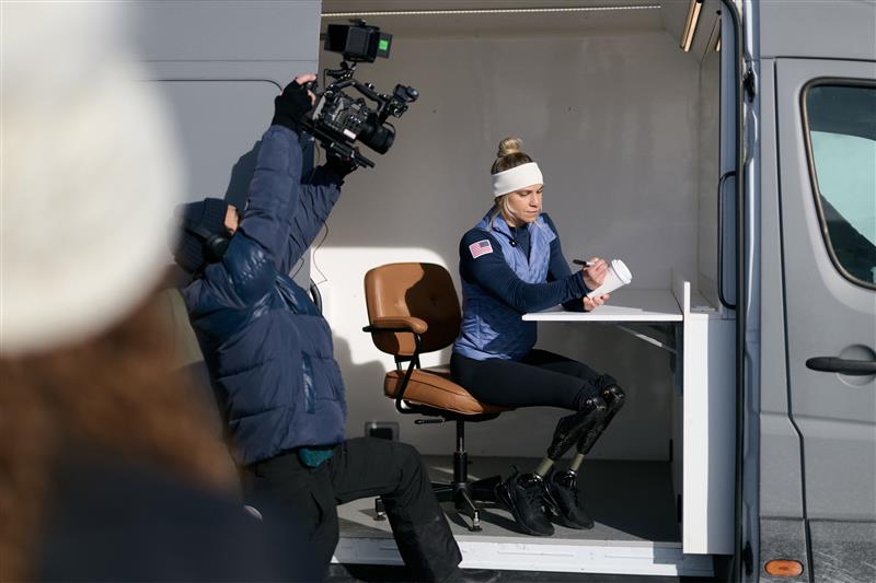 A woman with a headband sits at a desk in a mobile unit, writing on a notepad. A cameraman records her from the side.