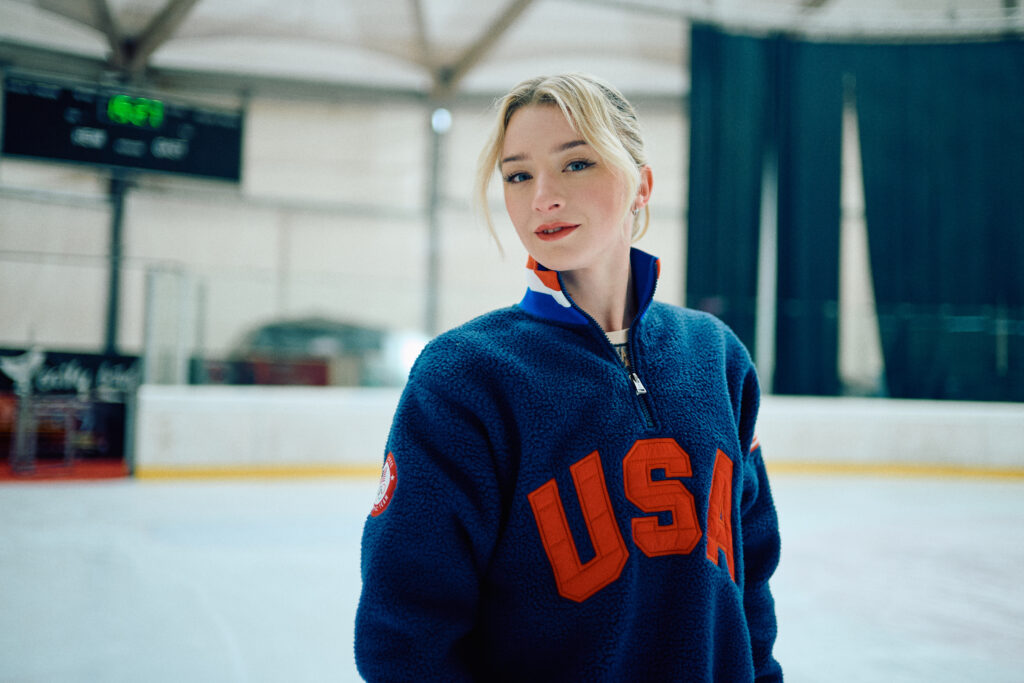A young woman stands on an ice rink, wearing a blue fleece jacket with "USA" prominently displayed in red. She has blonde hair styled elegantly and a confident expression. The background features an ice rink setting with soft lighting.