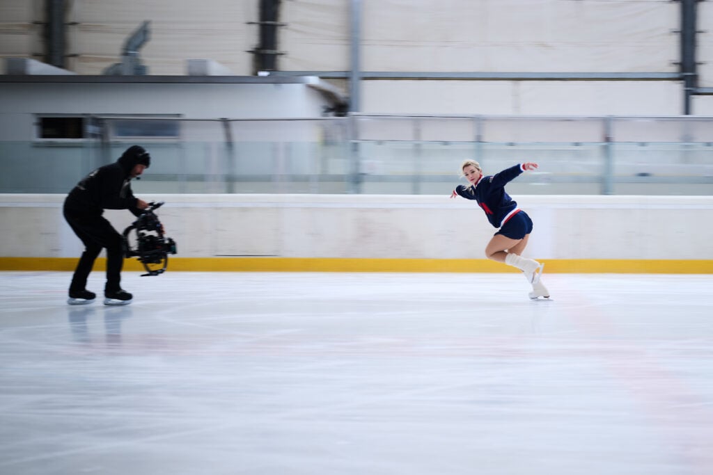 A figure skater in a blue outfit glides gracefully on the ice, while a cameraman captures the action. The scene conveys motion and elegance.