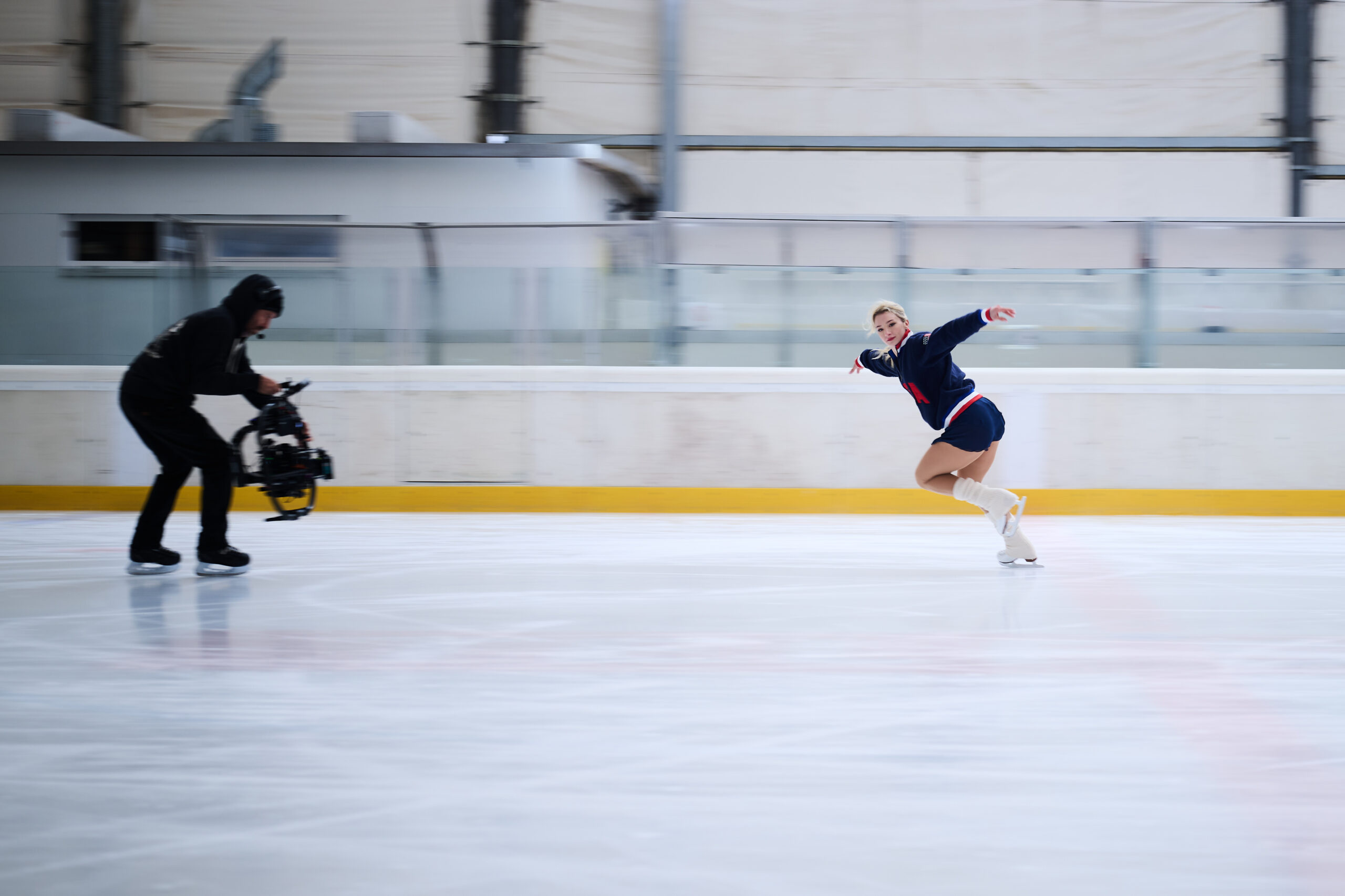 A figure skater in a blue outfit glides gracefully on the ice, while a cameraman captures the action. The scene conveys motion and elegance.