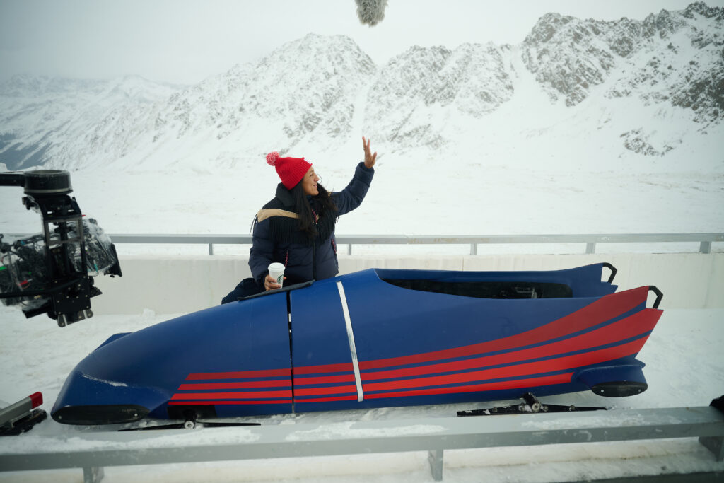 A woman in a red beanie and a puffy jacket sits in a blue bobsled, holding a coffee cup. Snowy mountains are visible in the background.