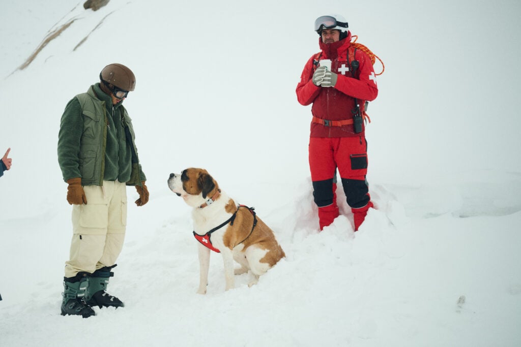 A Saint Bernard dog sits in the snow, wearing a harness. Two people are nearby: one in a green jacket and the other in a red rescue outfit, holding a cup.