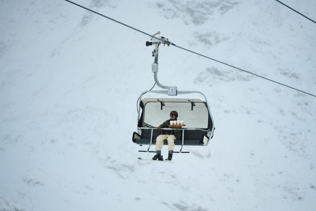 A skier sits on a chairlift, wearing a helmet and goggles, holding a tray with drinks. The snowy landscape surrounds them.