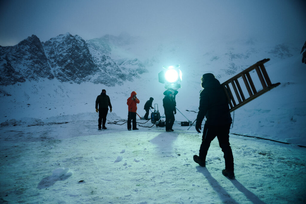 A snowy landscape with several crew members working. A large spotlight illuminates the scene, while one person carries a ladder.