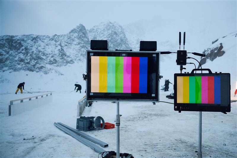 Two monitors displaying colorful test patterns are set up on stands in a snowy landscape, with workers in the background.
