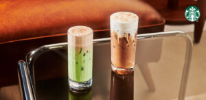 Two tall glasses of beverages are displayed on a glass table. The left glass contains a green drink with a frothy top, while the right glass features a layered iced coffee topped with foam and a sprinkle of cinnamon. A warm-toned background adds to the cozy ambiance.