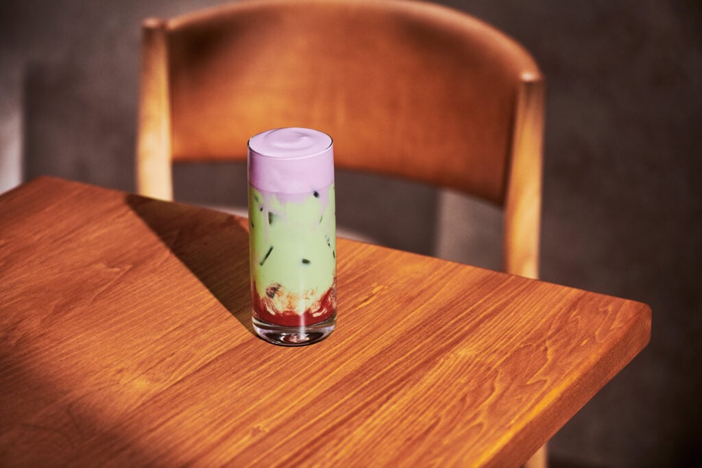 A tall glass filled with layered drinks: a purple frothy top, green liquid with ice, and a red base, placed on a wooden table.