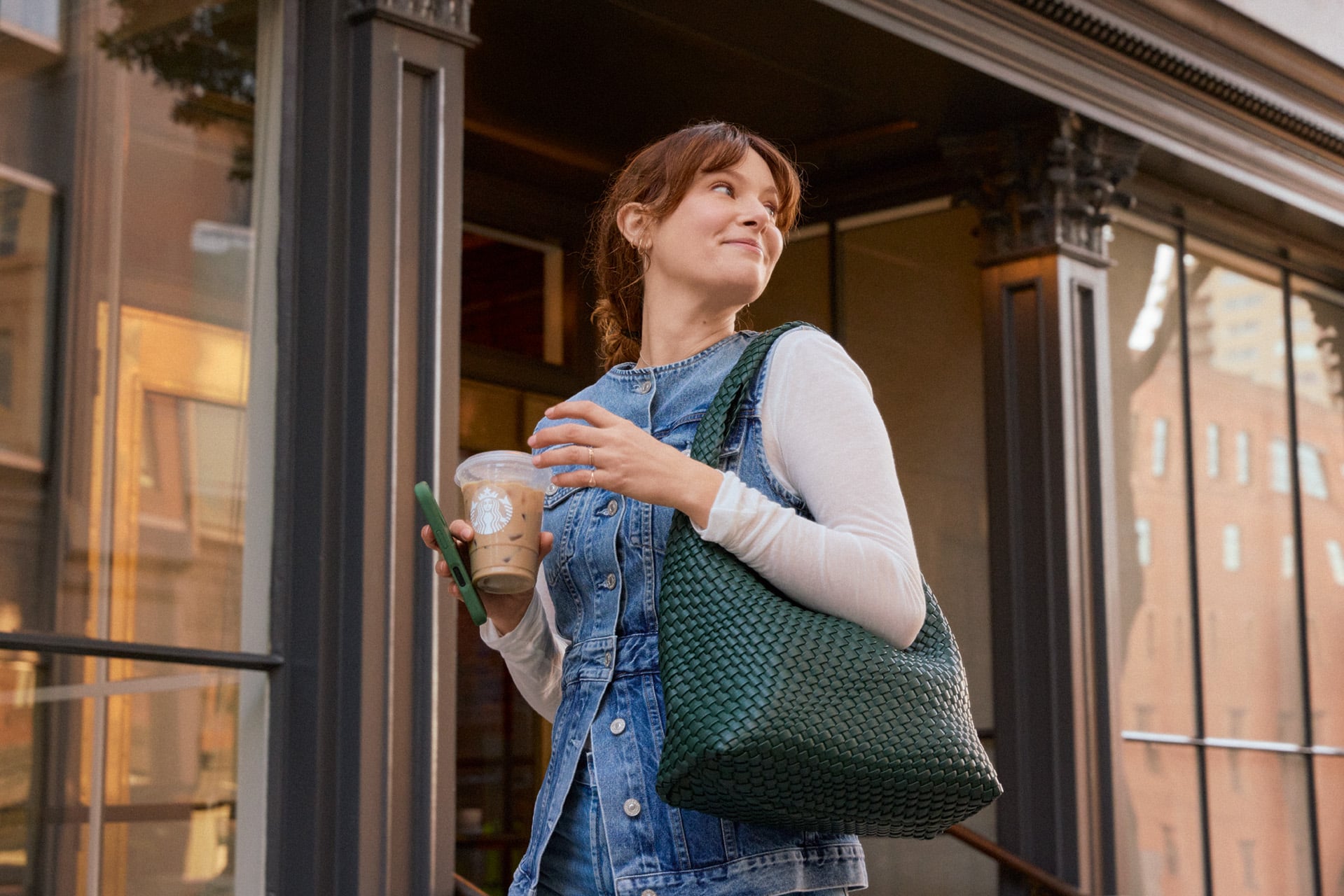 A woman stands outside a café, holding an iced coffee and a phone. She wears a denim dress over a white long-sleeve shirt and carries a large green woven bag.